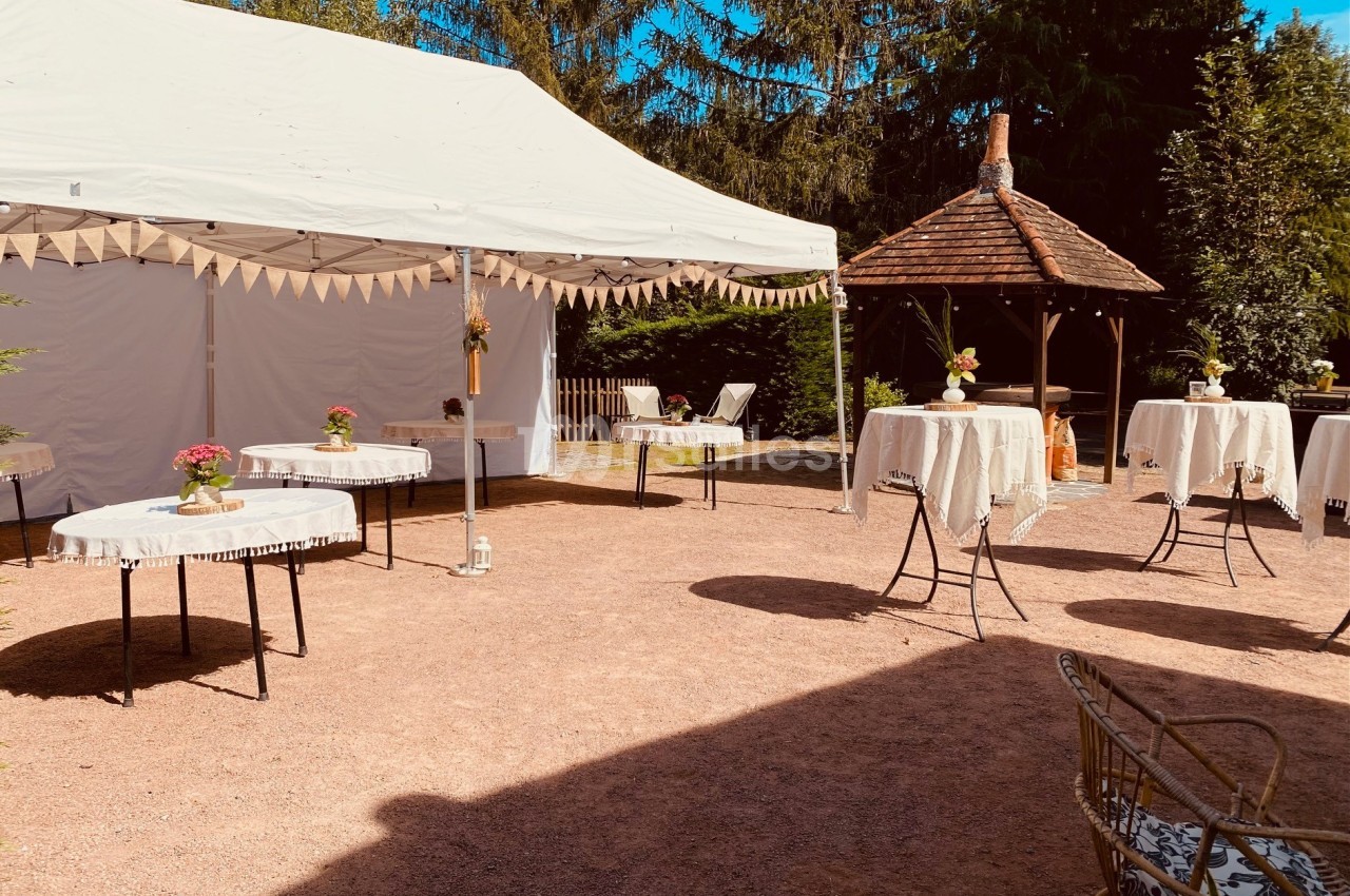 Espace extérieur aménagé pour un événement, avec tables décorées, tente blanche et kiosque en arrière-plan.