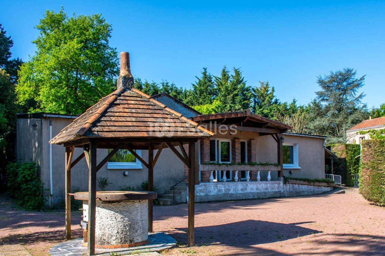 Maison avec façade beige, entourée de végétation, et un puits en pierre avec un toit en bois au premier plan.