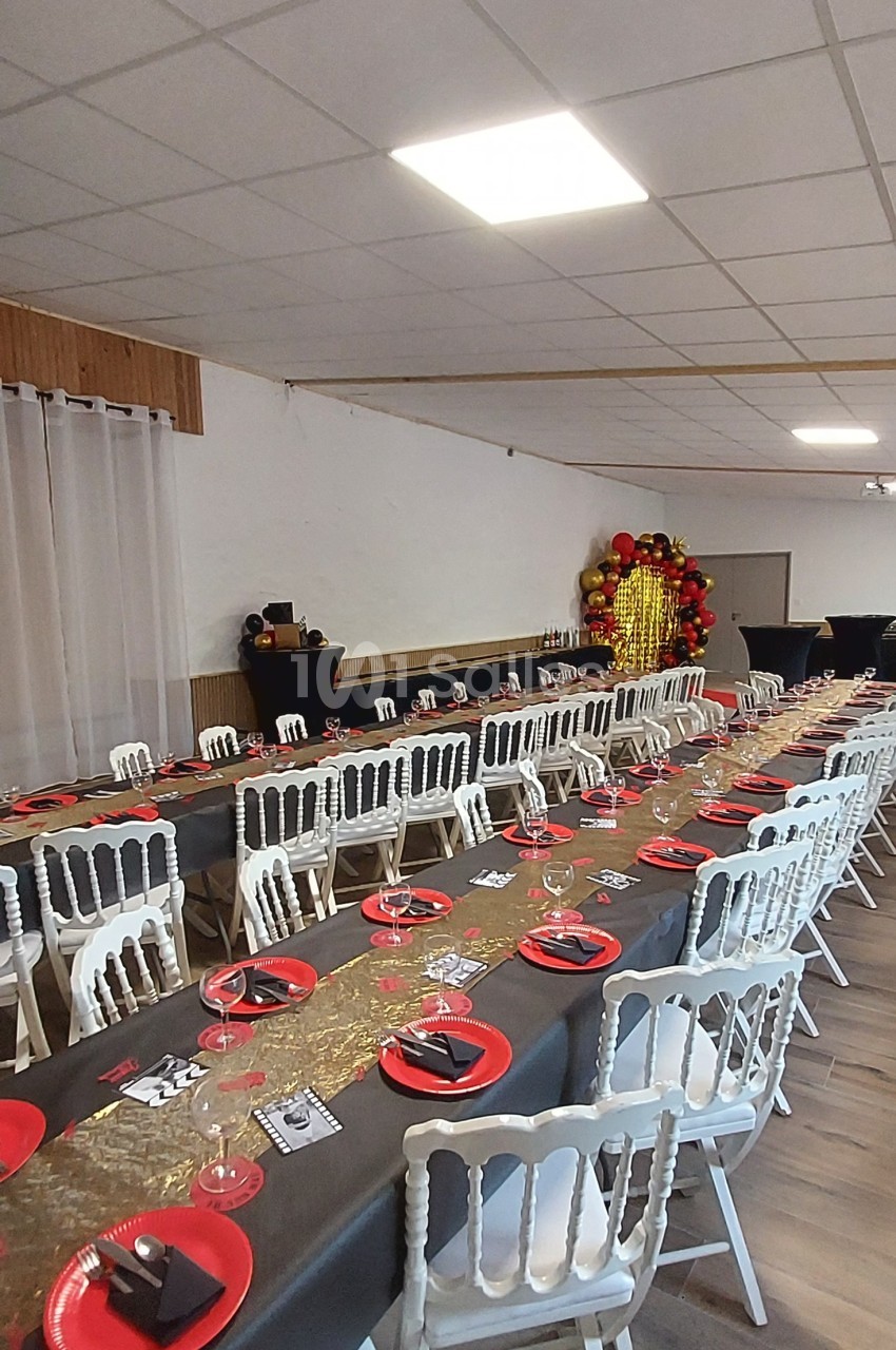 Salle décorée pour un événement avec longues tables dressées, nappes noires, assiettes rouges et décoration dorée en fond.