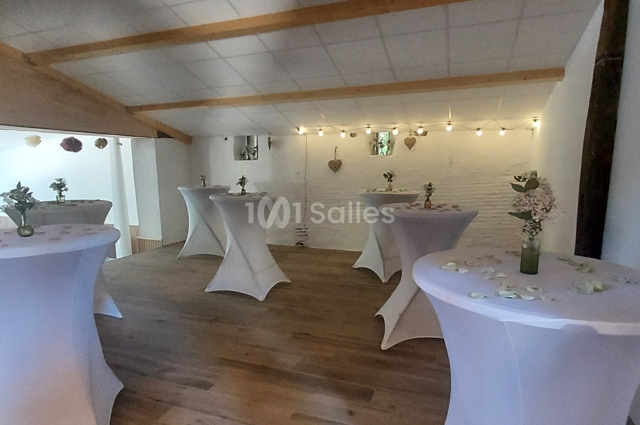 Salle décorée avec des tables hautes recouvertes de nappes blanches, ornées de fleurs et de pétales, éclairée par des…