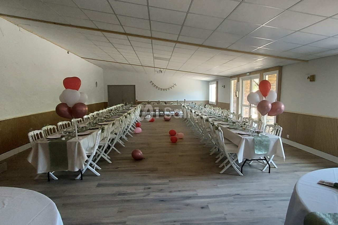 Salle décorée pour un événement avec des tables alignées, des ballons colorés et de la lumière naturelle.
