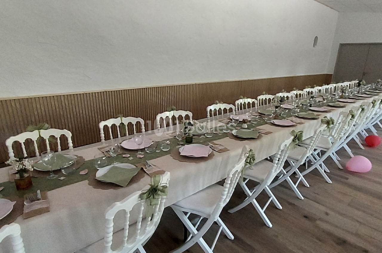 Salle décorée pour un événement avec longues tables dressées, nappes beiges, assiettes colorées et ballons au sol.