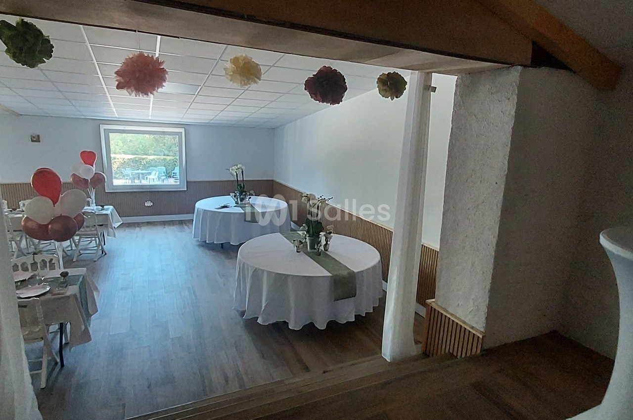 Salle décorée pour un événement, avec tables rondes dressées, ballons et guirlandes suspendues.