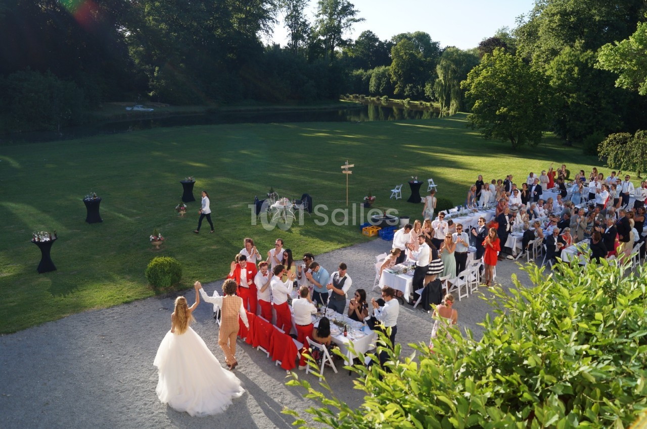 Un couple de mariés salue des invités assis en extérieur, dans un jardin verdoyant par une journée ensoleillée.