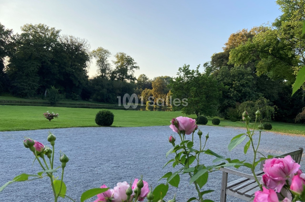 Allée gravillonnée bordée de roses avec vue sur une pelouse, des arbres et un plan d'eau sous un ciel dégagé.