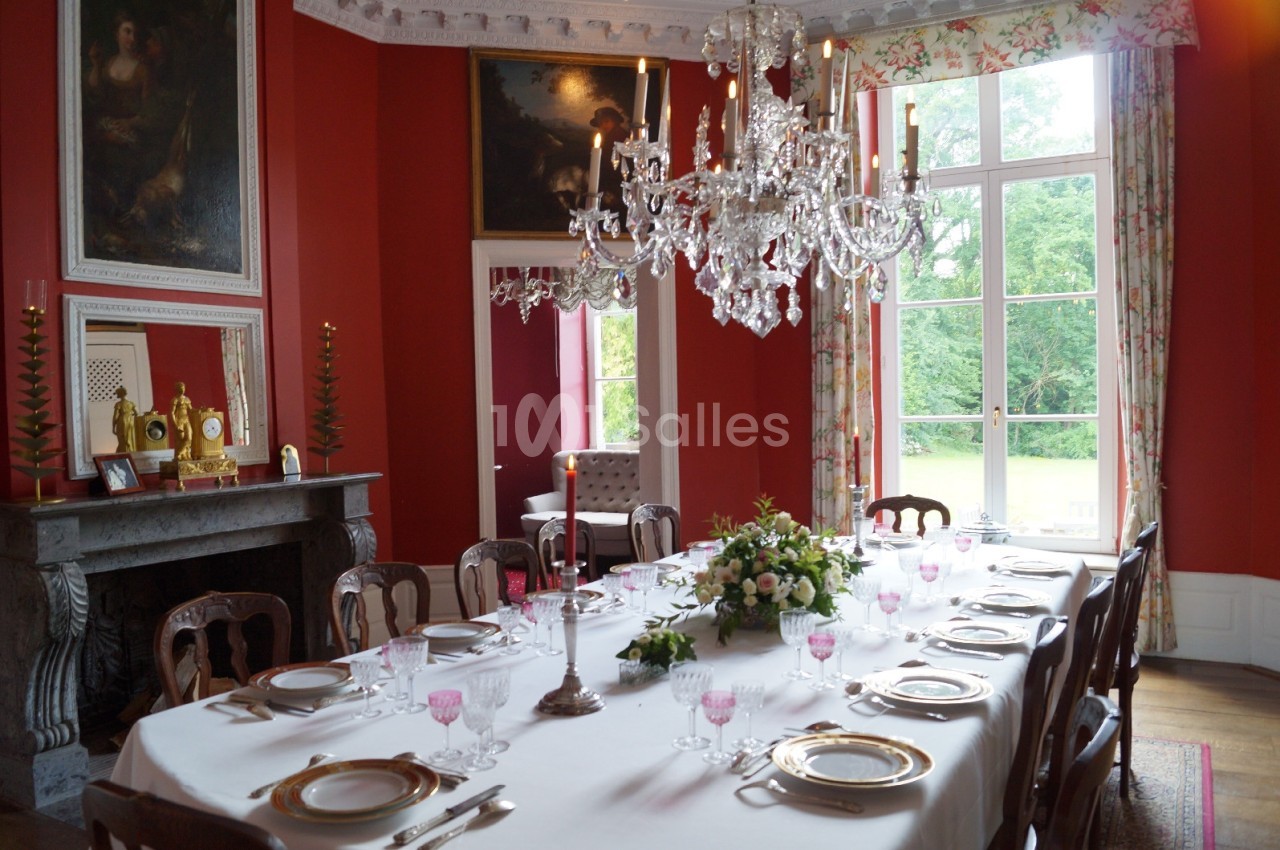 Salle à manger élégante avec table dressée, chandelier central, murs rouges et vue sur un jardin par de grandes fenêtres.