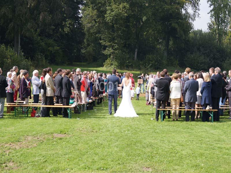 Un couple en tenue de mariage avance entre des invités assis à l'extérieur, dans un parc verdoyant.