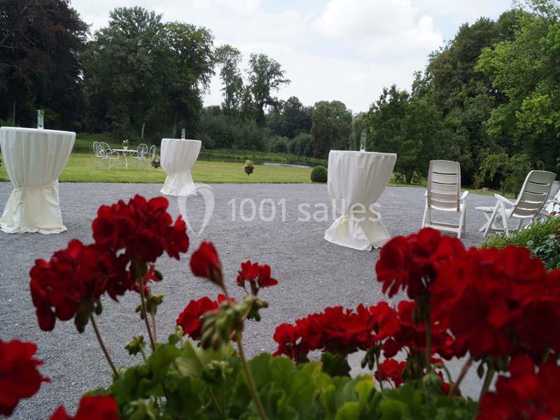 Jardin avec tables hautes drapées de blanc, chaises blanches et géraniums rouges au premier plan.
