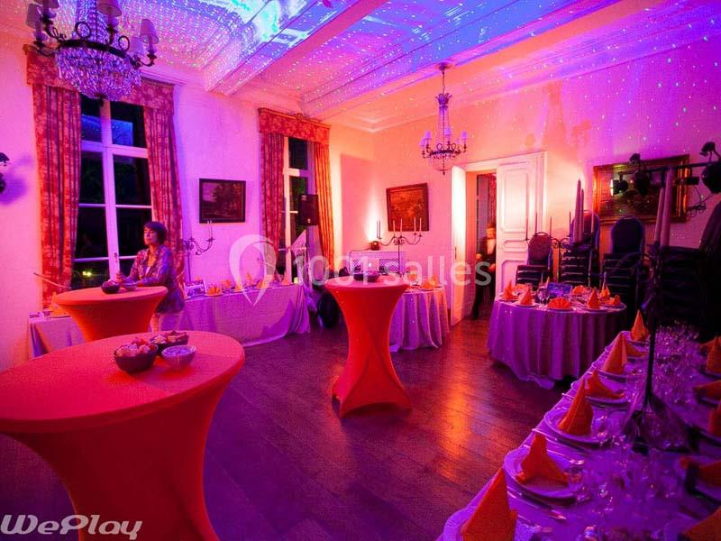 Salle décorée pour un événement, avec tables dressées, nappes colorées et éclairage d'ambiance violet.