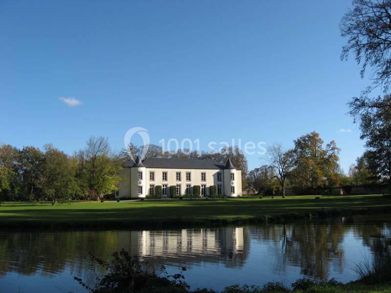 Manoir blanc entouré d'arbres, avec une pelouse et un étang reflétant le bâtiment sous un ciel bleu.