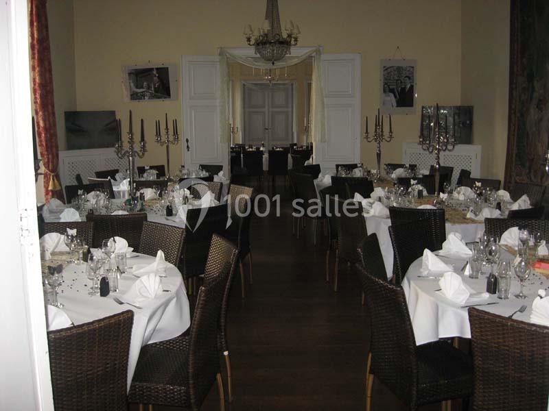Salle de réception élégamment décorée avec des tables rondes, nappes blanches, chaises en osier et chandeliers.