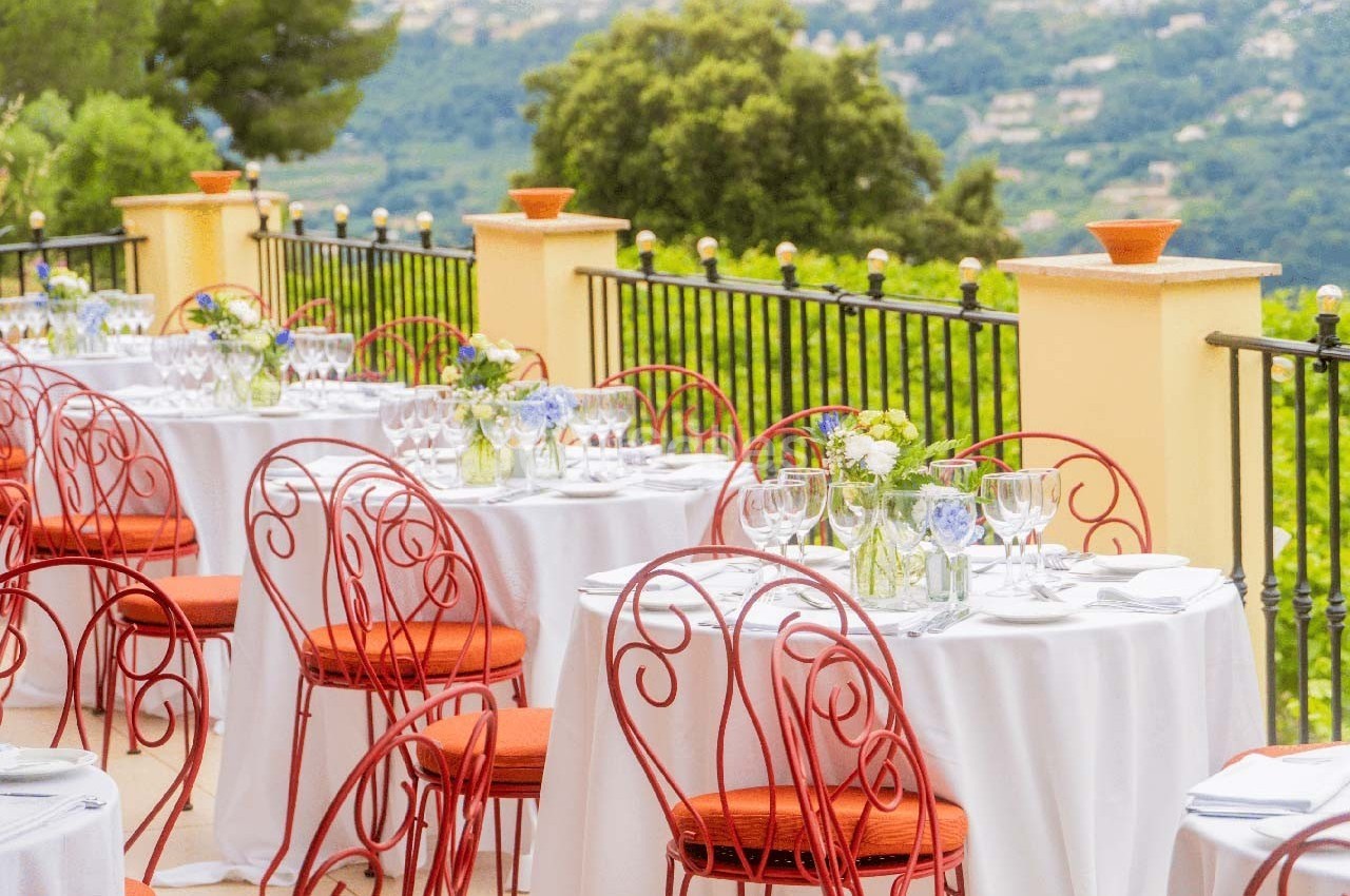 Tables dressées avec nappes blanches et chaises rouges sur une terrasse surplombant un paysage verdoyant.