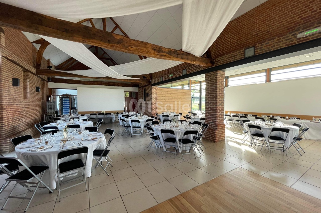 Salle de réception lumineuse avec tables rondes dressées, chaises noires et poutres apparentes en bois.