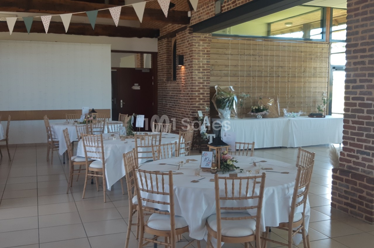 Salle de réception décorée avec des tables rondes, nappes blanches, chaises en bois clair et guirlandes suspendues.