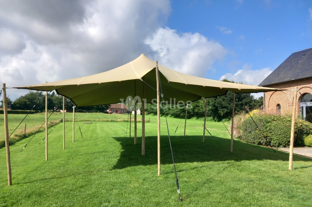 Tente beige montée sur une pelouse, soutenue par des poteaux en bois, avec un bâtiment en briques à droite.