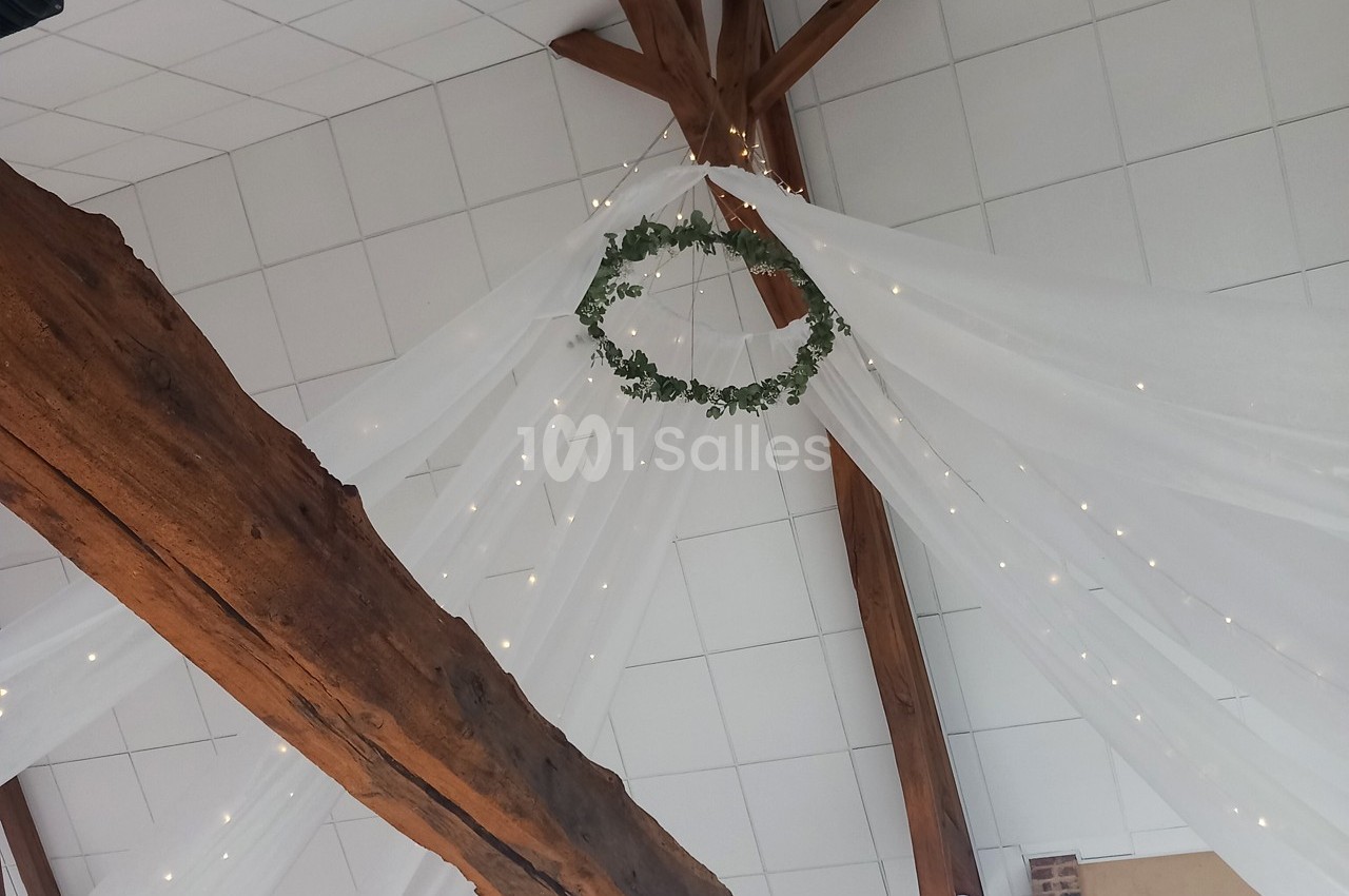 Guirlande lumineuse et couronne végétale suspendues sous une charpente en bois avec drapés blancs.