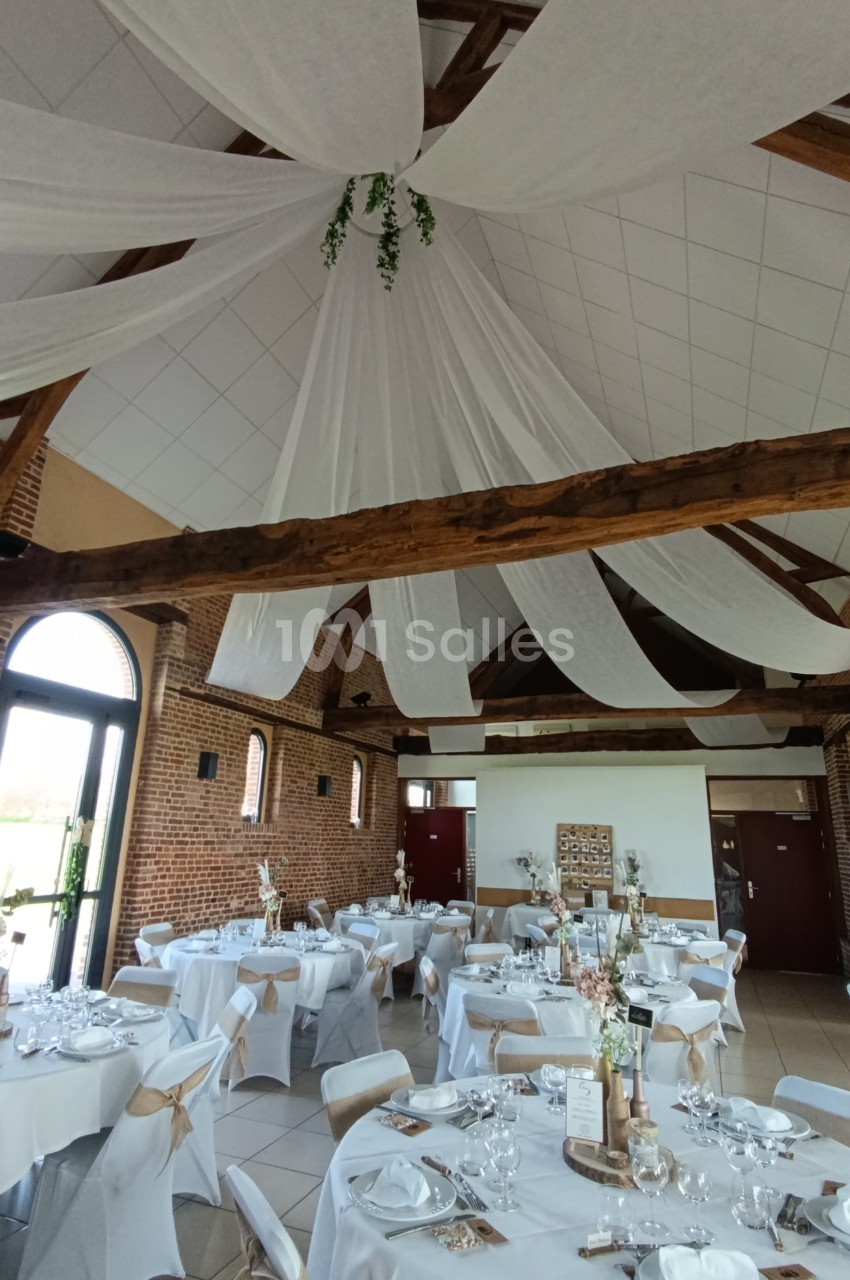 Salle de réception décorée avec des tables rondes dressées, nappes blanches et voilages suspendus au plafond.