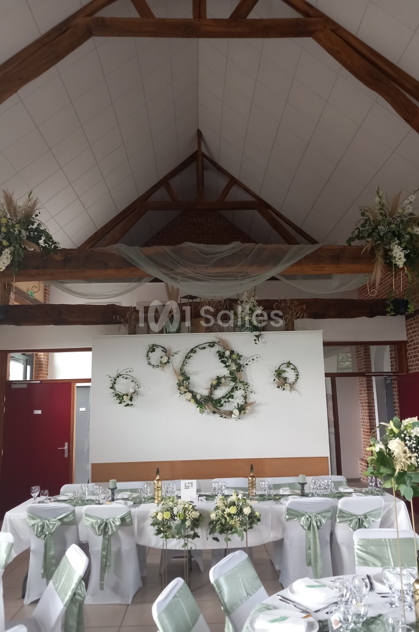 Salle de réception décorée pour un mariage, avec des tables dressées, des chaises ornées de rubans verts et des fleurs…