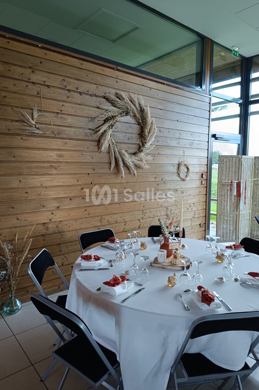 Table ronde dressée pour un repas, décorée de serviettes rouges, avec un mur en bois orné de couronnes de fleurs séchées.
