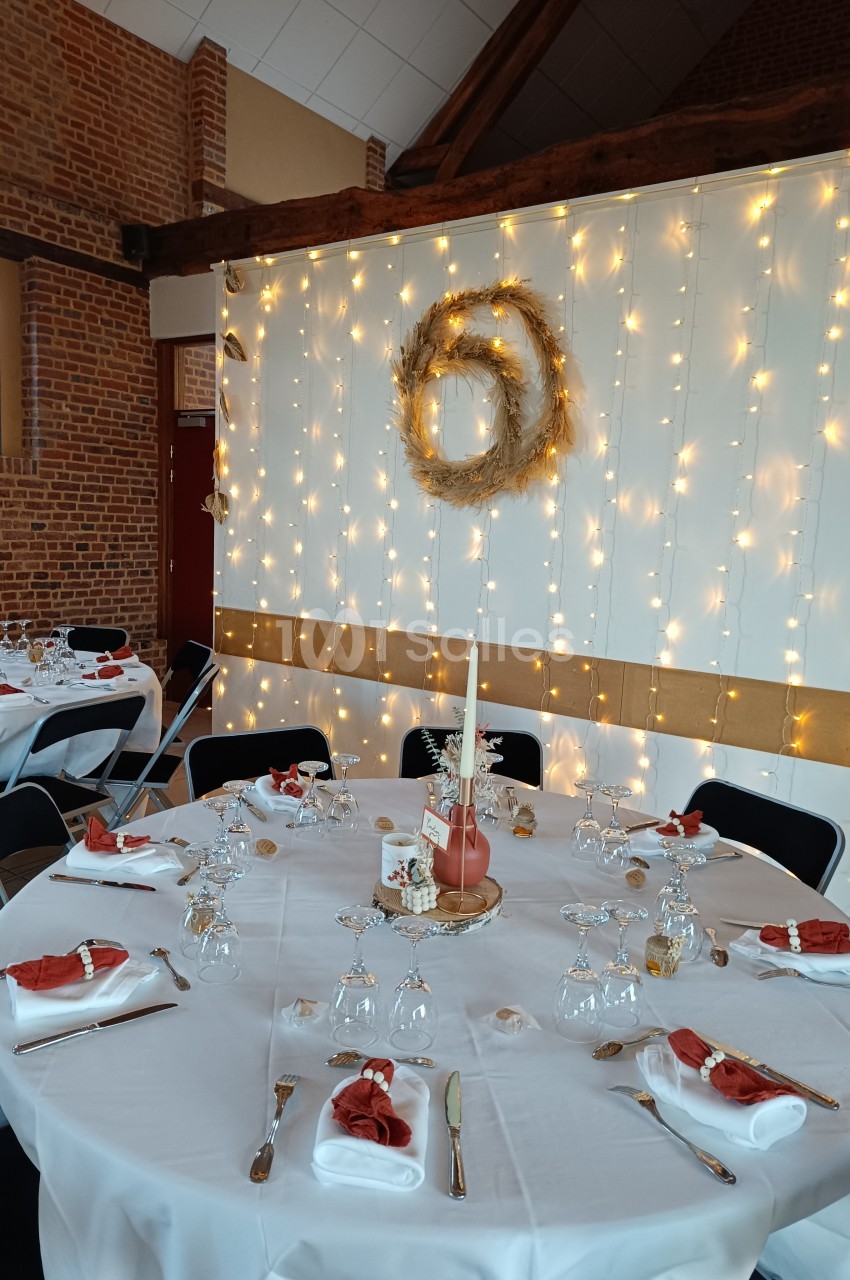 Salle décorée pour un événement avec tables dressées, nappes blanches, serviettes rouges et guirlandes lumineuses sur un mur.