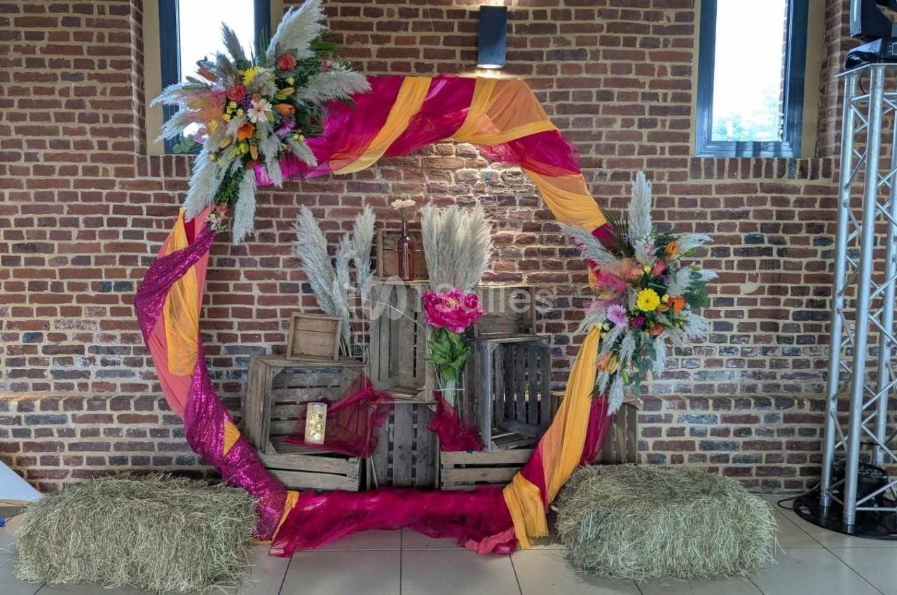 Cercle décoratif avec tissus colorés, fleurs et bottes de paille, devant un mur de briques dans un espace intérieur.