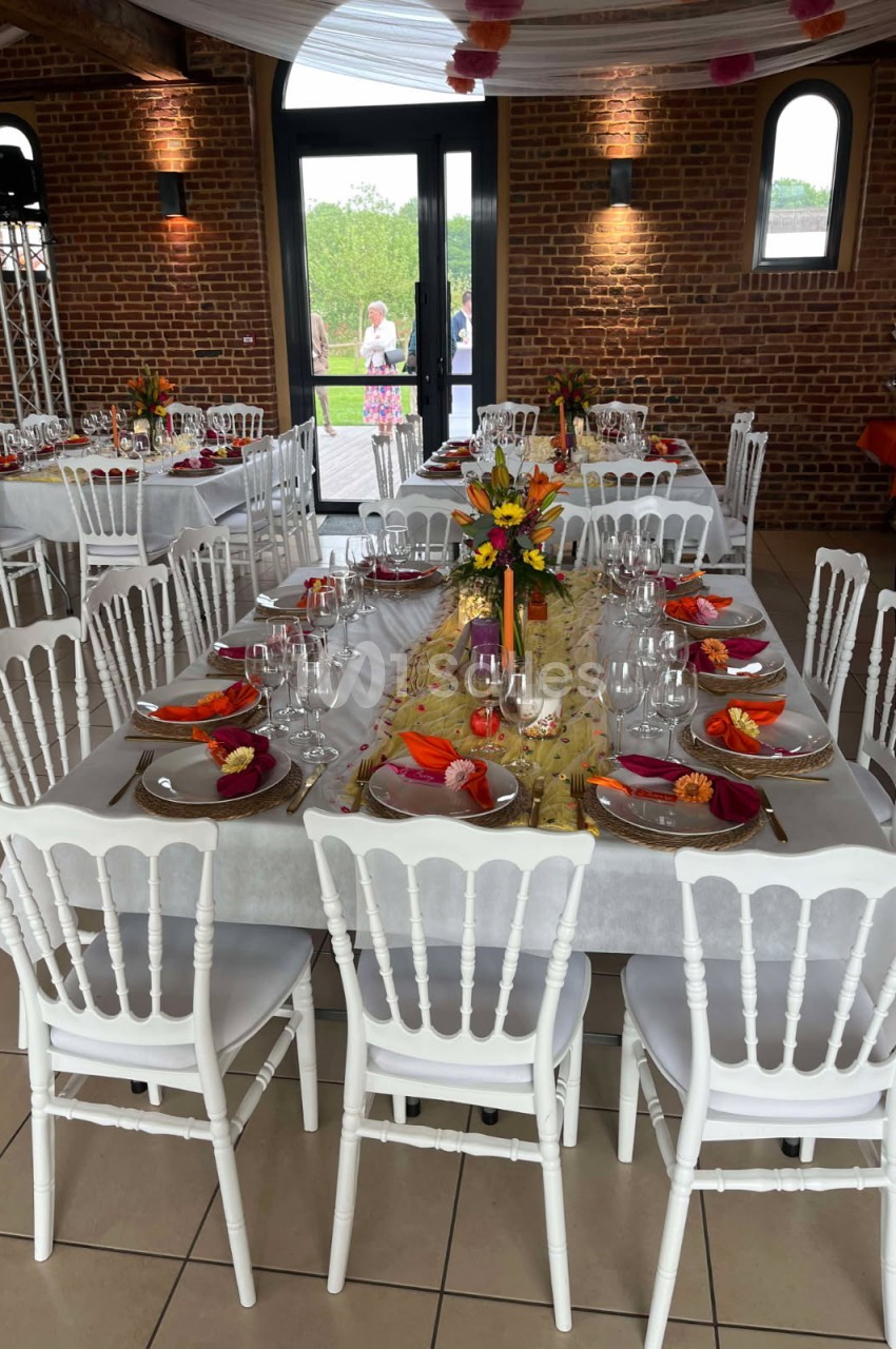 Salle décorée pour un repas festif avec tables dressées, nappes blanches, chaises blanches et compositions florales colorées.