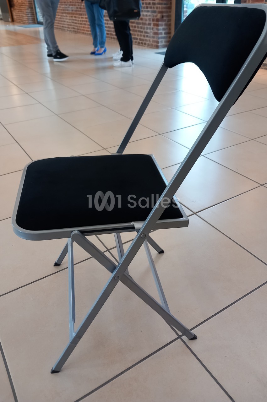 Chaise pliante noire avec structure métallique argentée, posée sur un sol carrelé beige dans un espace intérieur.