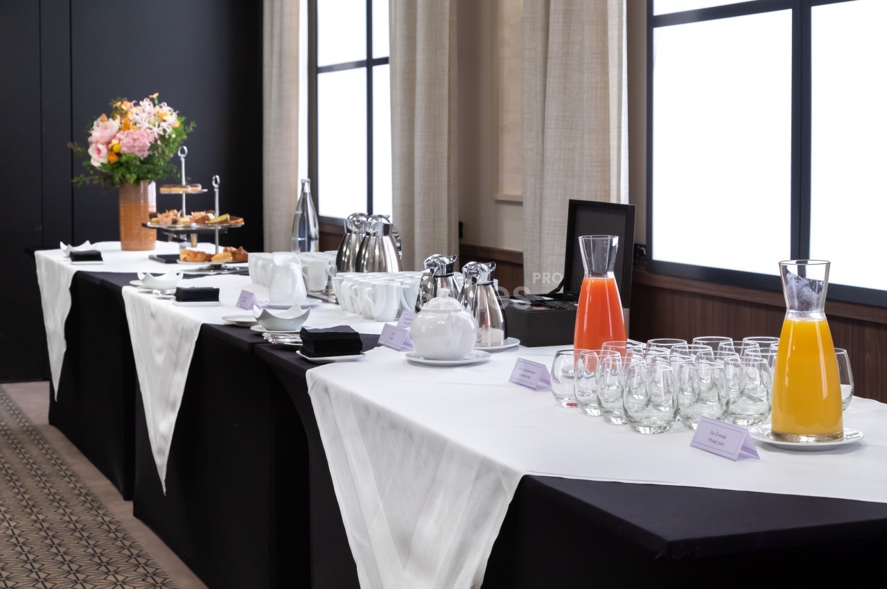 Table de buffet avec carafes de jus, tasses, assiettes, viennoiseries et un bouquet de fleurs en arrière-plan.