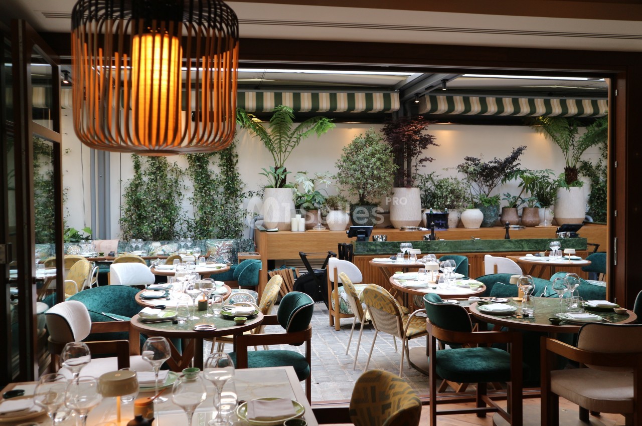 Salle de restaurant élégante avec tables dressées, chaises colorées et terrasse végétalisée en arrière-plan.