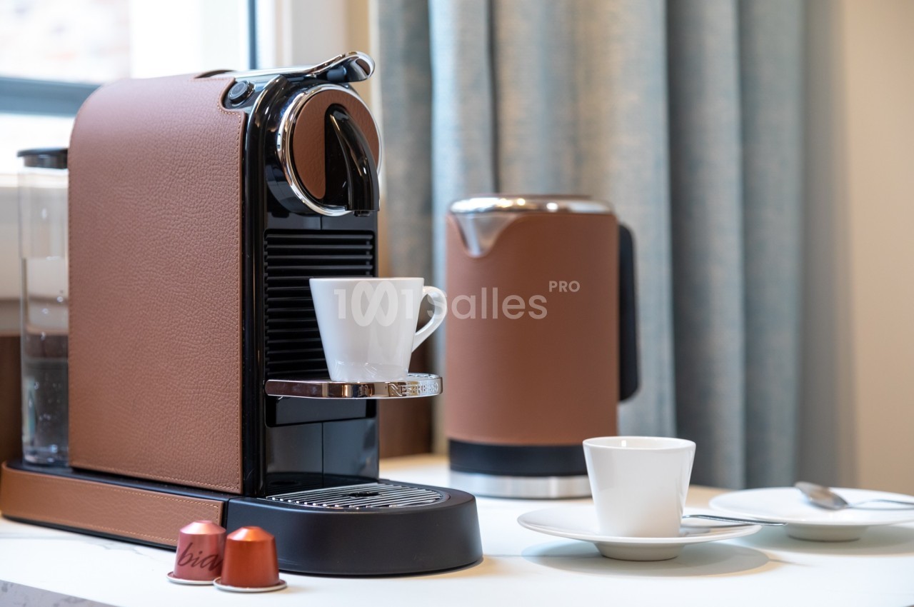 Machine à café avec une tasse blanche posée sur le support, capsules et accessoires visibles sur un plan de travail.