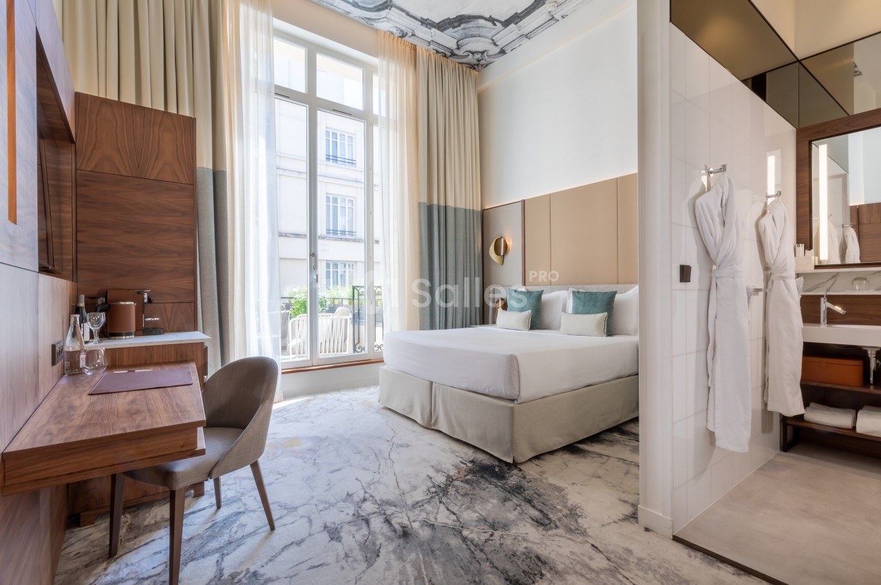 Chambre lumineuse avec un lit double, un bureau en bois, des peignoirs accrochés et une grande fenêtre donnant sur l…