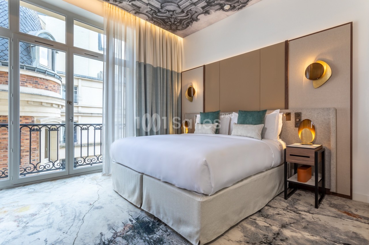 Chambre d'hôtel lumineuse avec un grand lit, des lampes murales dorées, un plafond décoré et une vue sur des bâtiments…