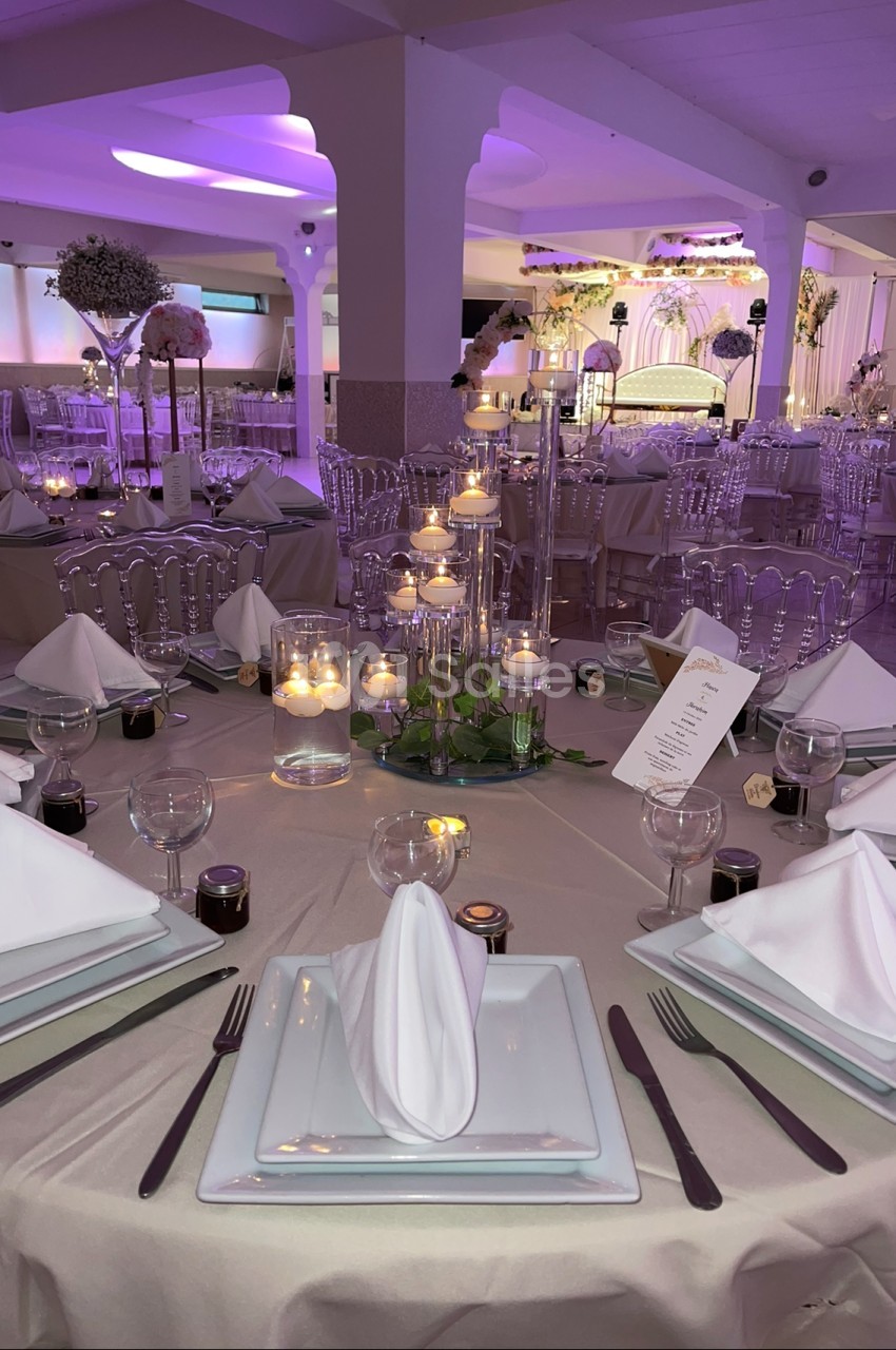 Salle de réception décorée avec des tables élégantes, nappes beiges, bougies et éclairage violet tamisé.