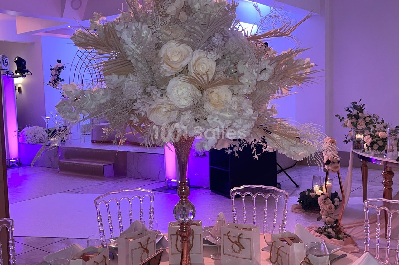 Centre de table floral avec roses blanches et feuillage beige, dans une salle décorée de lumières violettes.