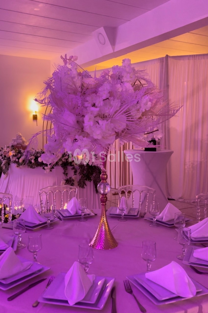 Centre de table floral avec des fleurs blanches et roses sur une table dressée dans une salle de réception éclairée.