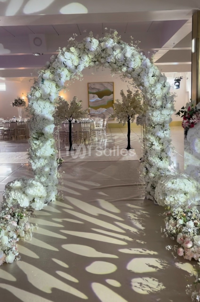 Arche de mariage décorée de fleurs blanches, entourée de chaises transparentes et d'arbres artificiels.
