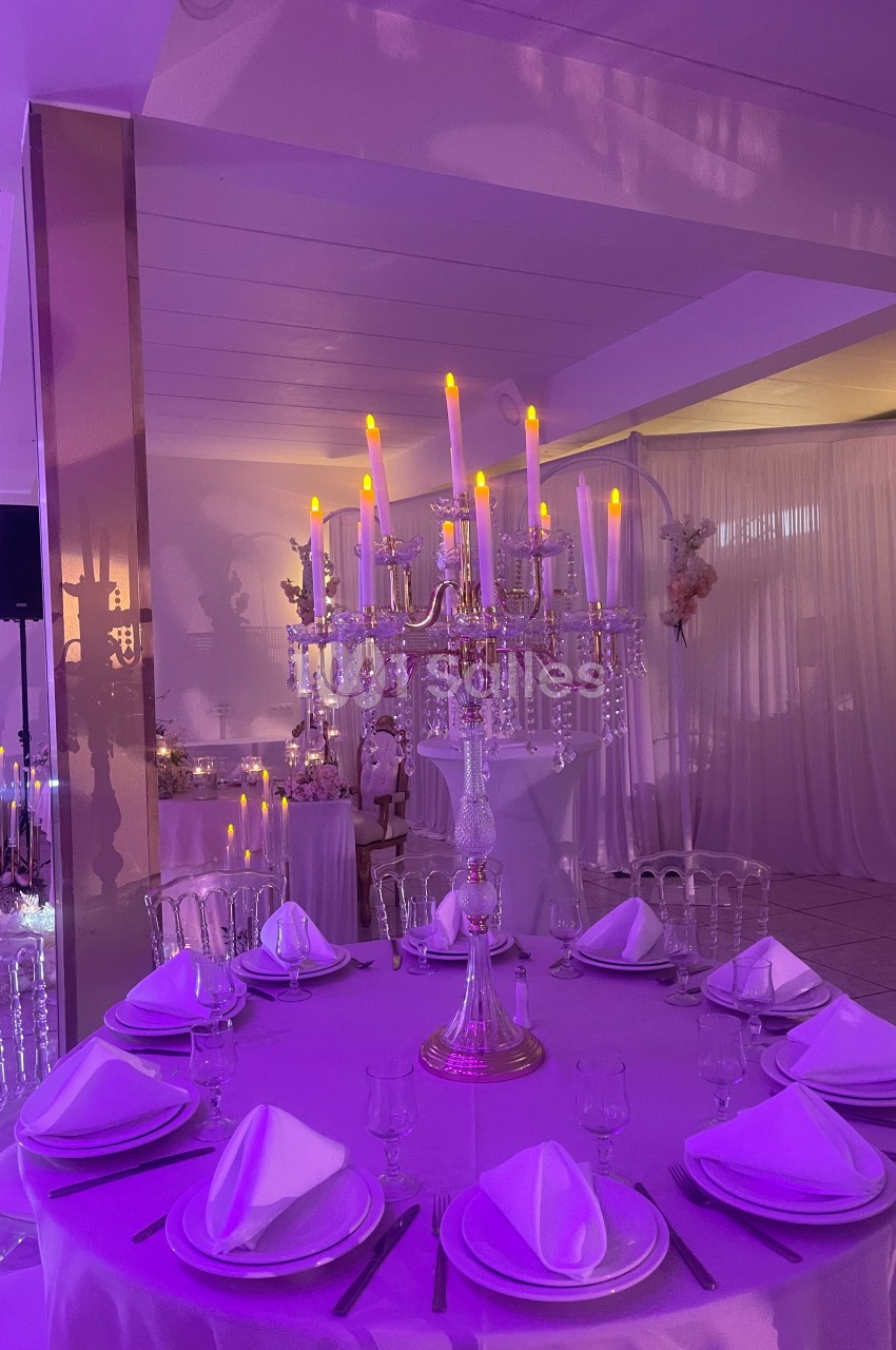 Centre de table avec chandelier doré et bougies LED, entouré de tables dressées dans une salle éclairée en violet.