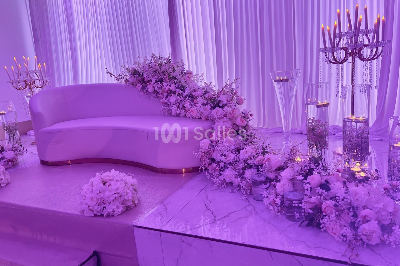 Canapé blanc décoré de fleurs roses et blanches, entouré de chandeliers et de rideaux violets dans un décor élégant.