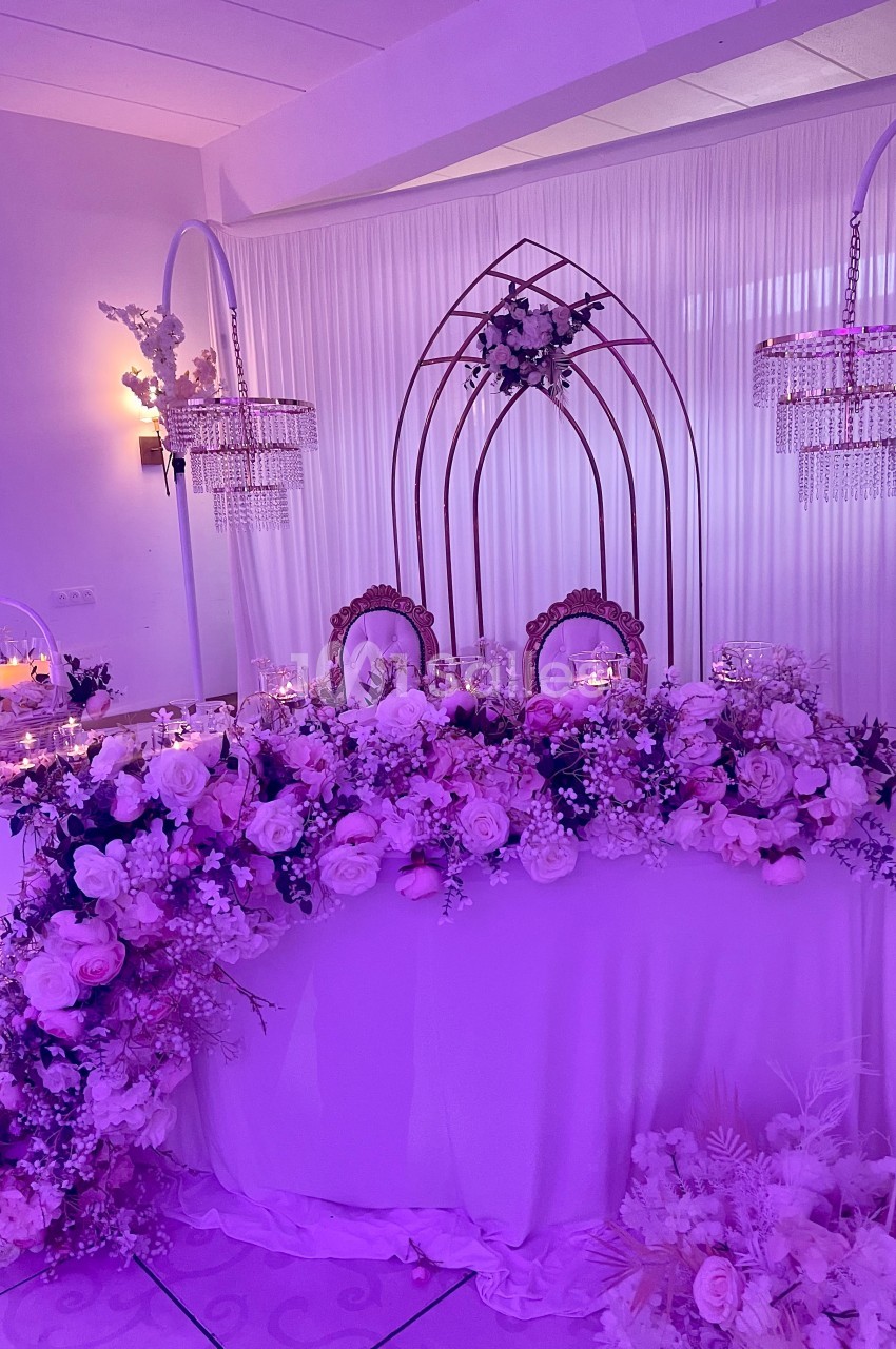 Table ornée de fleurs roses et blanches, décorée pour un événement, avec arche métallique et éclairage tamisé.