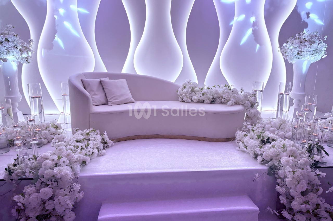 Canapé blanc entouré de fleurs blanches et de bougies, placé sur une estrade devant un décor lumineux ondulé.