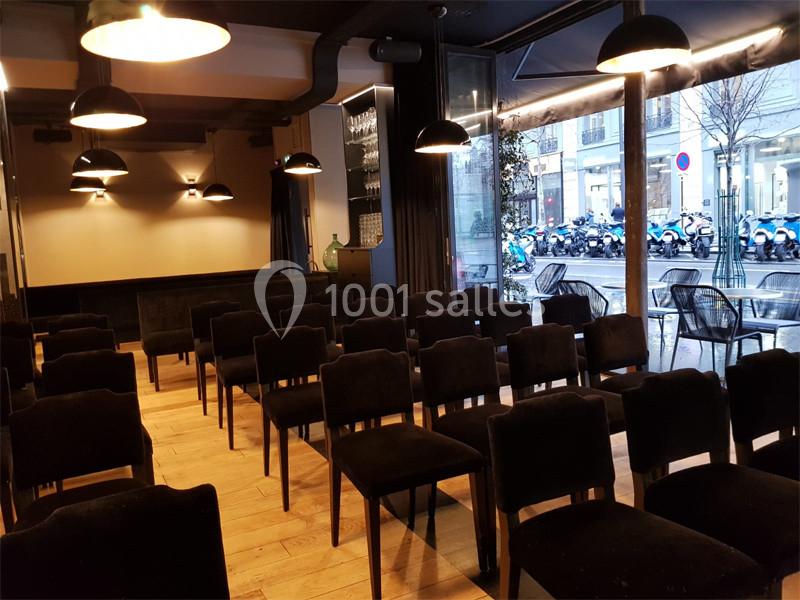 Salle aménagée avec des rangées de chaises noires, éclairée par des suspensions, donnant sur une rue avec des vitrines.