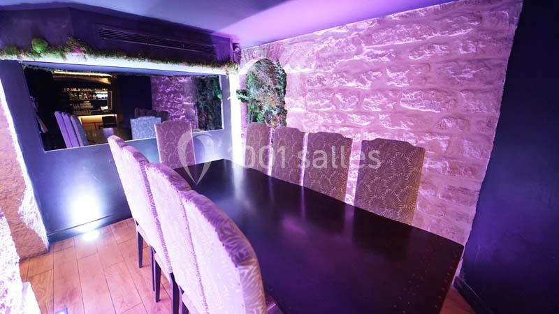 Salle à manger avec table en bois sombre, chaises rembourrées et murs en pierre éclairés par une lumière violette.