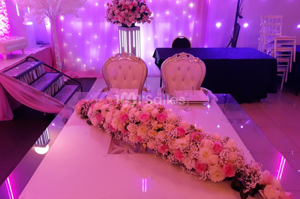 Table décorée pour un mariage avec un chemin de fleurs roses et blanches, deux chaises élégantes et un fond lumineux.