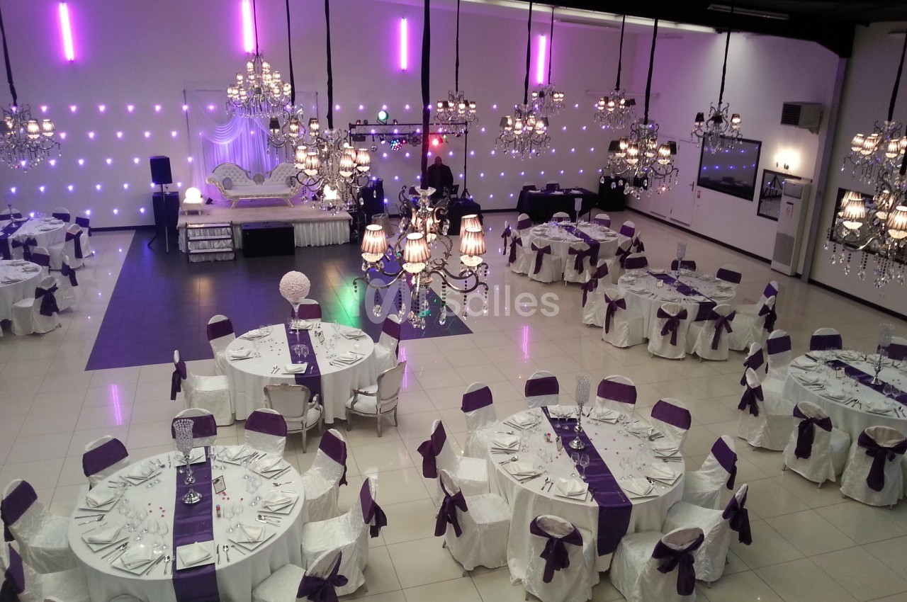 Salle de réception décorée avec des nappes blanches et des accents violets, éclairée par des lustres et des lumières murales.
