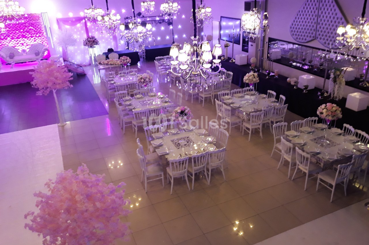 Salle de réception décorée avec des tables dressées, des chaises blanches, des lustres et des éclairages roses.