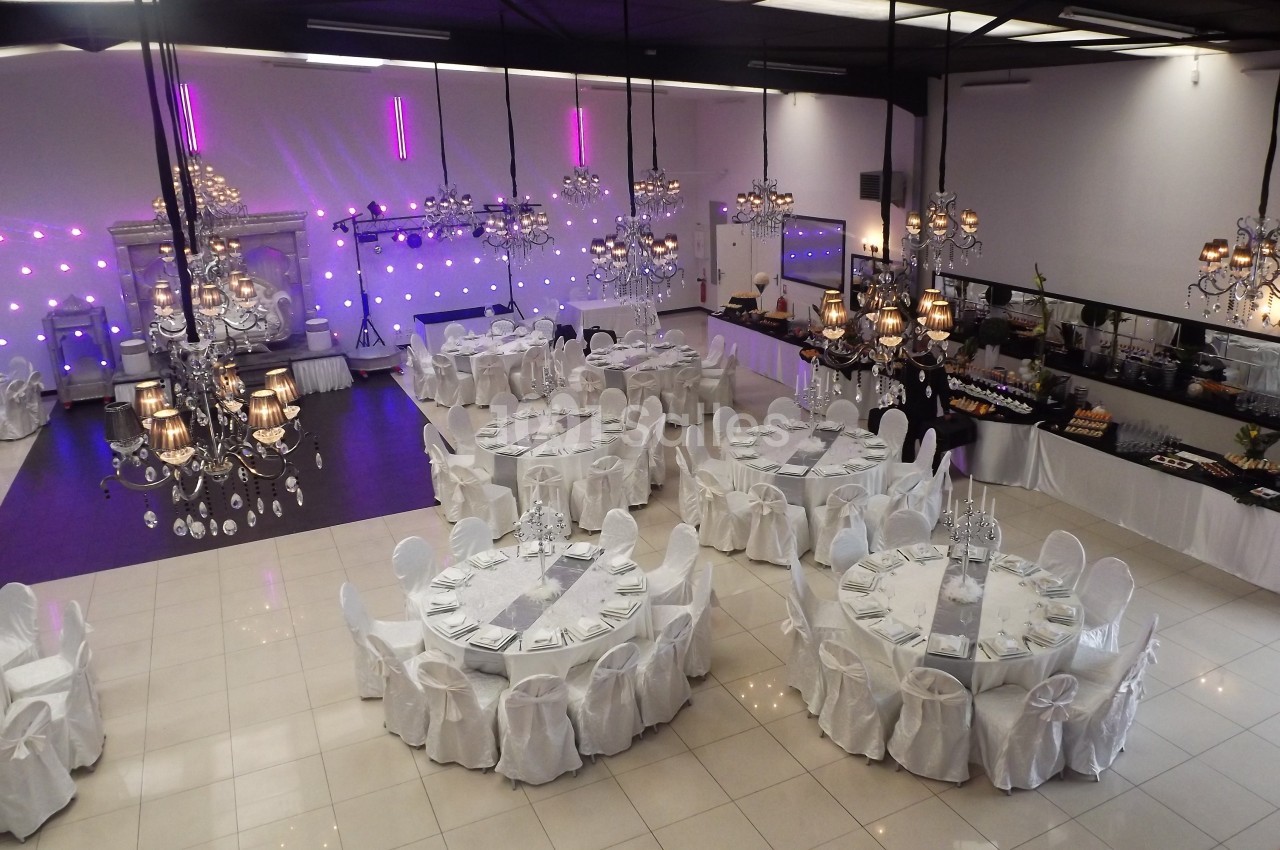 Salle de réception élégante avec tables rondes dressées, chaises blanches et lustres suspendus.