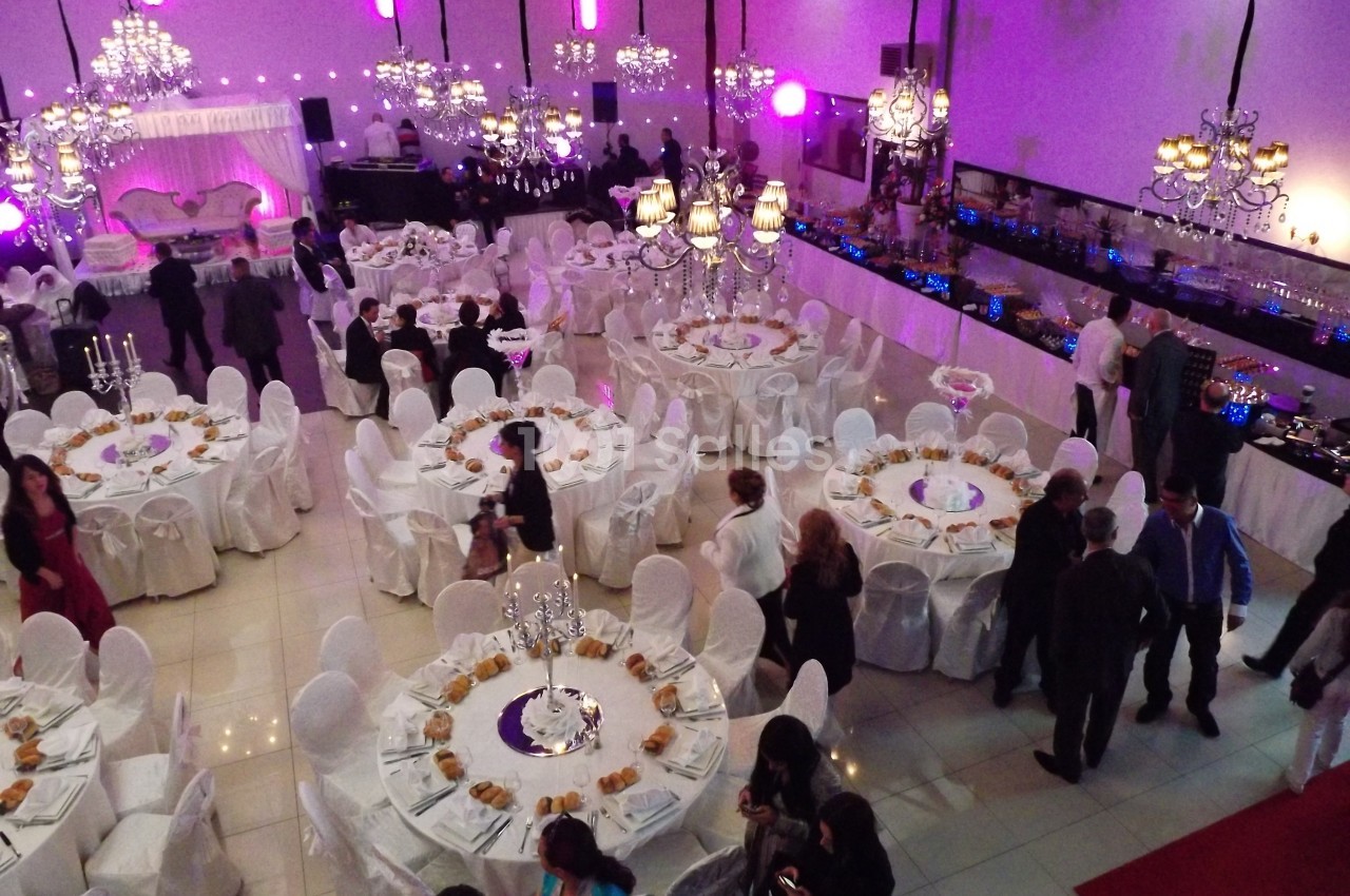 Salle de réception décorée avec des tables rondes dressées, des chaises blanches et des lustres suspendus.