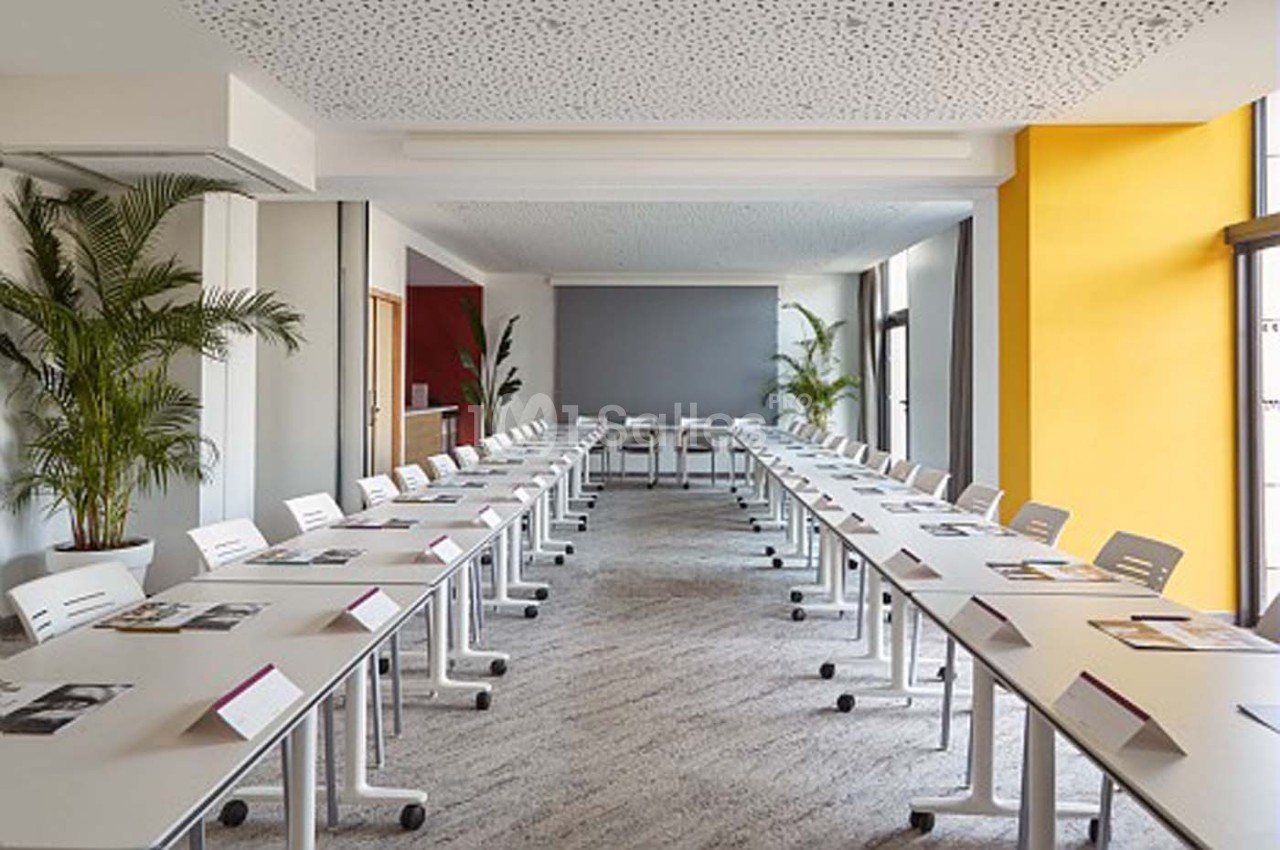 Salle de réunion lumineuse avec tables blanches disposées en U, chaises modernes et plantes en arrière-plan.