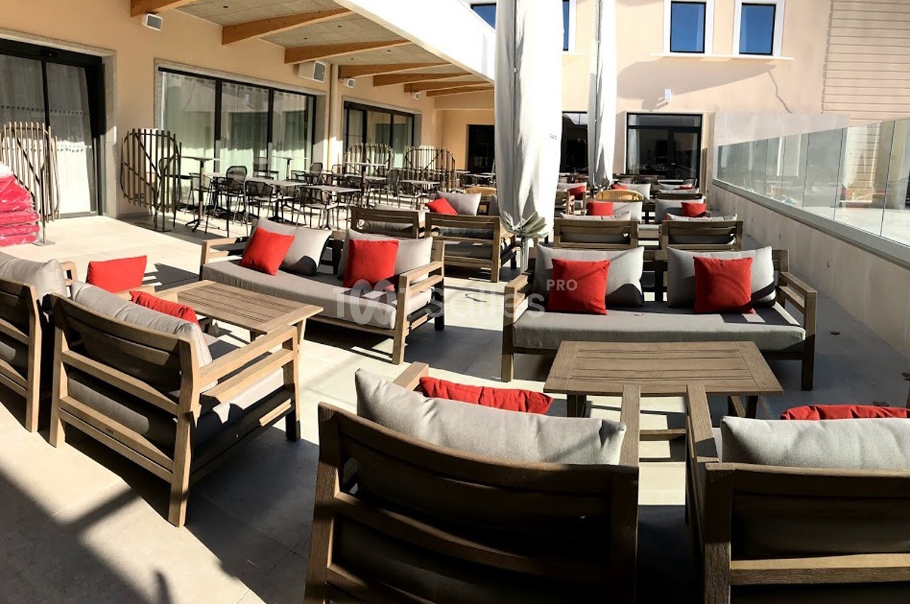 Terrasse ensoleillée avec mobilier en bois, coussins rouges et gris, tables basses et baies vitrées en arrière-plan.