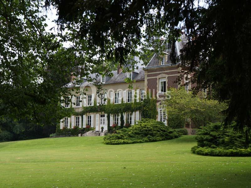 Grande maison en briques et pierres entourée d'un jardin verdoyant avec pelouse et arbres.